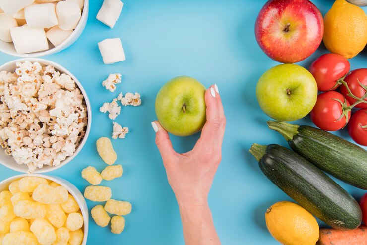 Imagem contendo alimentos saudáveis de um lado e alimentos não tão saudáveis do outro lado. Do lado esquerdo: pipoca, salgadinho e marshmallow. Do lado direito: maçã, tomate, abobrinha e no centro da imagem uma maçã verdade.